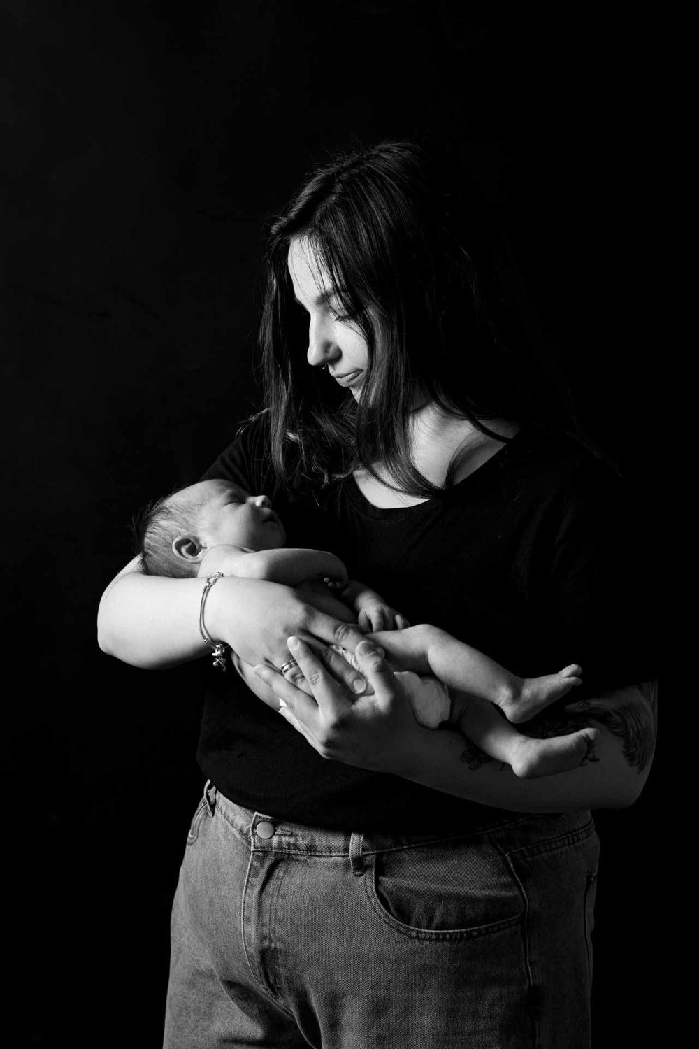A tender black and white portrait of a mother cradling her newborn baby against a dark backdrop.