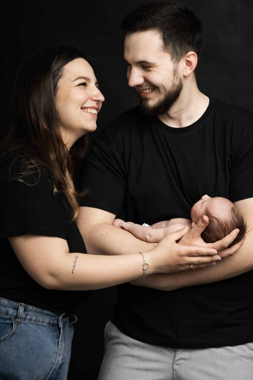 A couple in black shirts share an intimate moment while holding their newborn baby against a dark background.