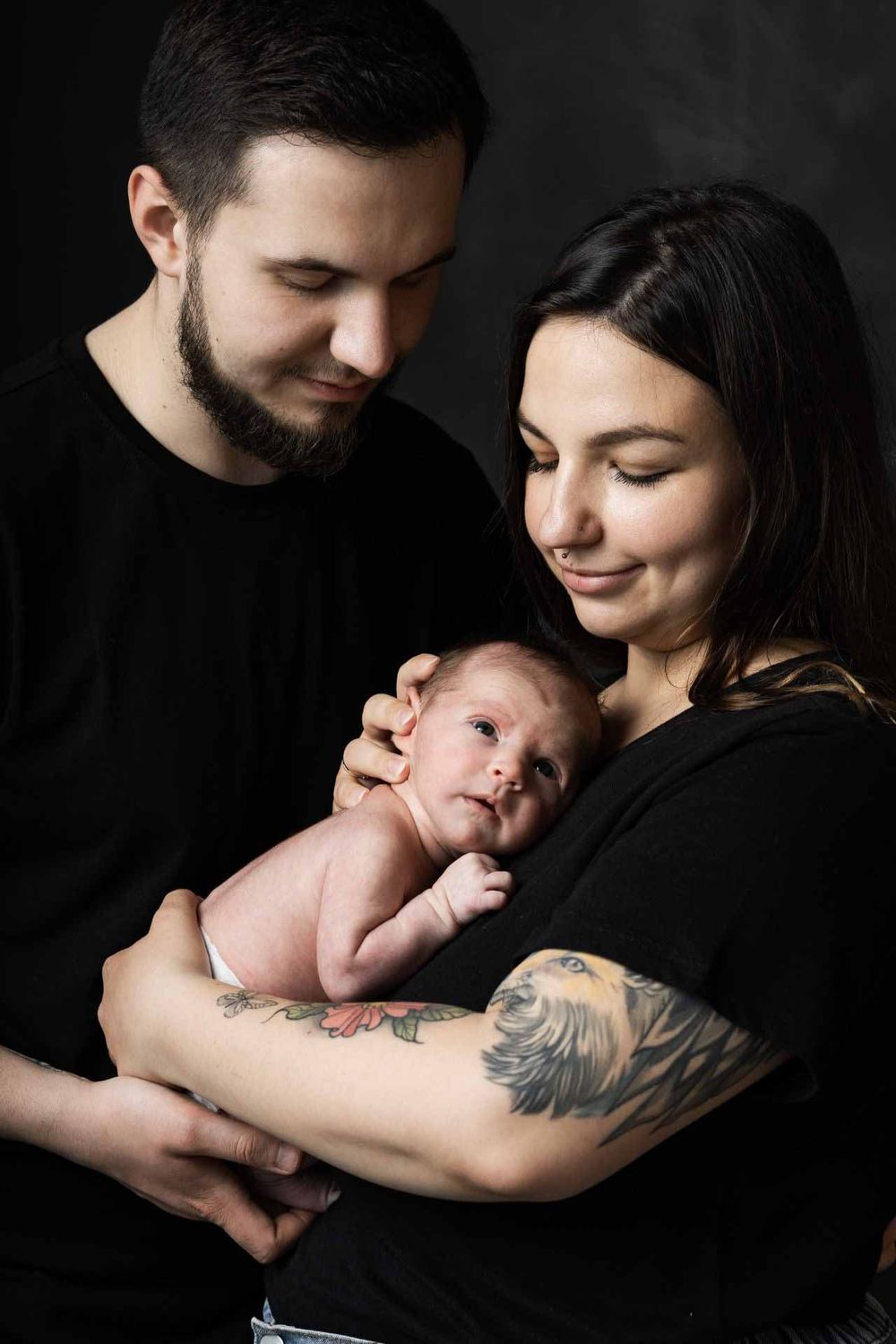 Parents in dark clothing share a loving moment with their newborn baby against a black background.