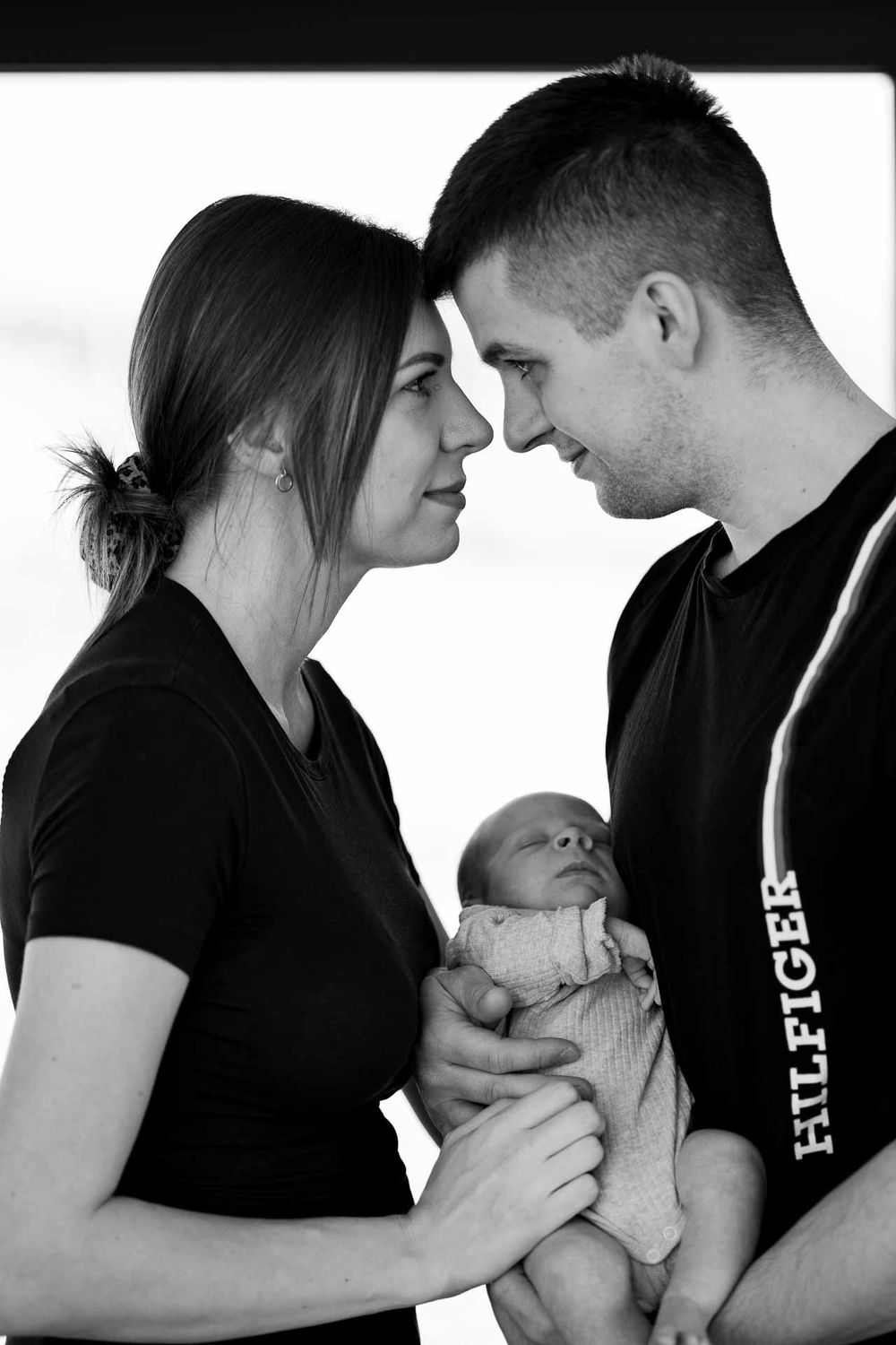 A black and white portrait of parents sharing an intimate moment while holding their sleeping newborn.