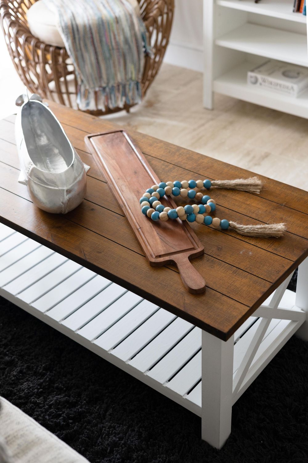 Farmhouse coffee table with dark wood top, white base, decorative cutting board, blue bead garland, and wicker chair in background.