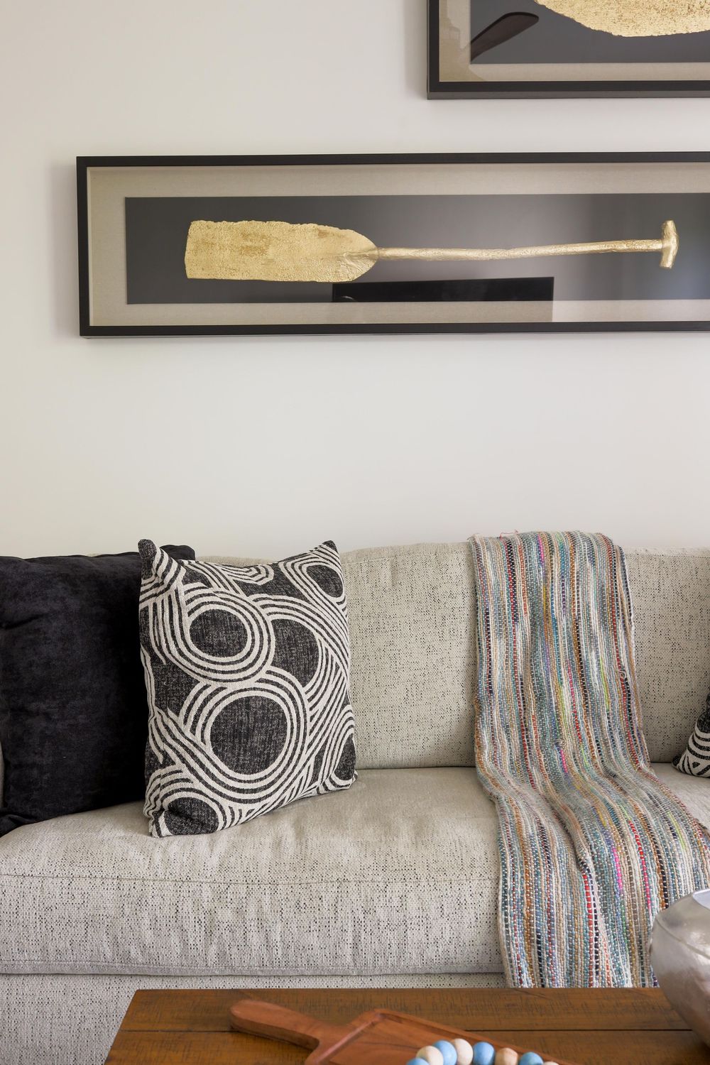 Gray sofa with black and white patterned pillows, colorful throw blanket, and framed golden oar wall art above.