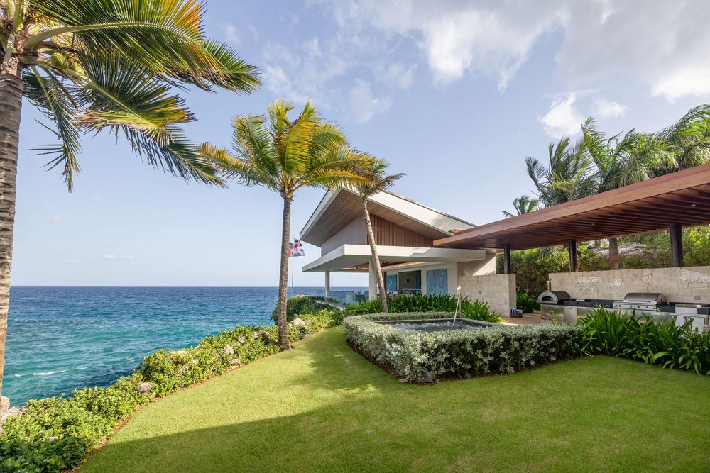 Luxury oceanfront villa with lush green lawn, palm trees, and stunning turquoise sea views under a partly cloudy sky.