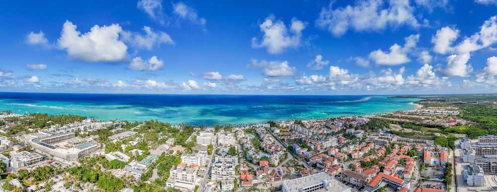 Aerial panoramic view of a coastal city with turquoise waters, white sand beaches, and urban development under blue skies. Bavaro Real Este