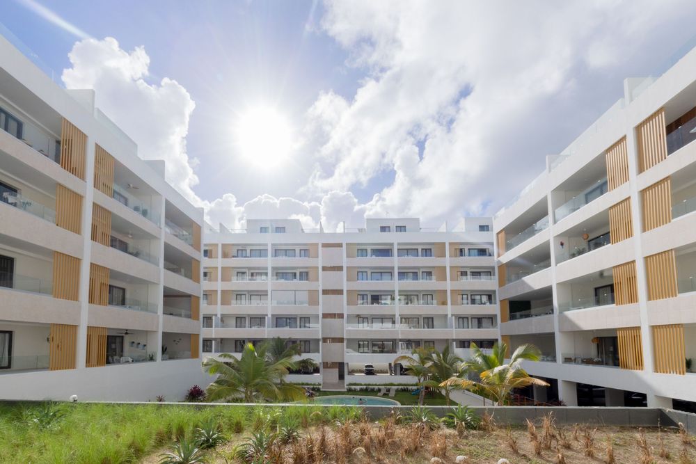 Modern apartment complex with beige and white exterior featuring balconies and landscaped courtyard under bright sunlit sky.
