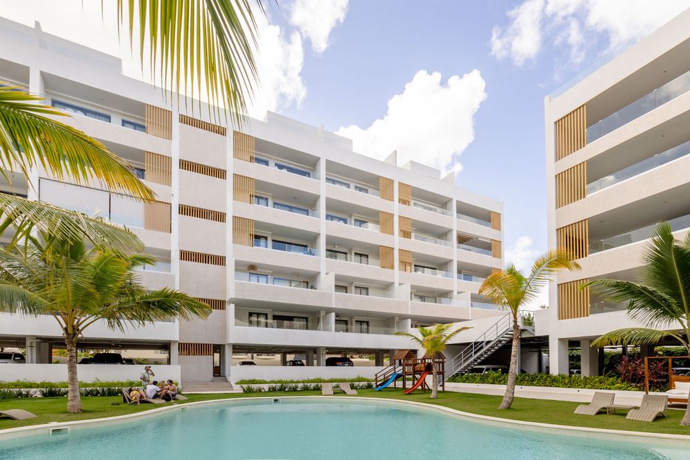 Luxury beachfront resort with white buildings, palm trees, and a pristine swimming pool overlooking the ocean. Bavaro