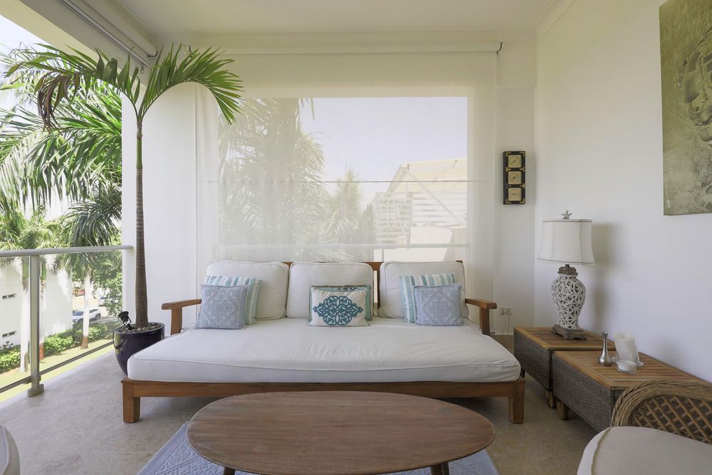 Bright tropical patio with a wooden daybed, blue accent pillows, round coffee table, and lush palm trees outside.
