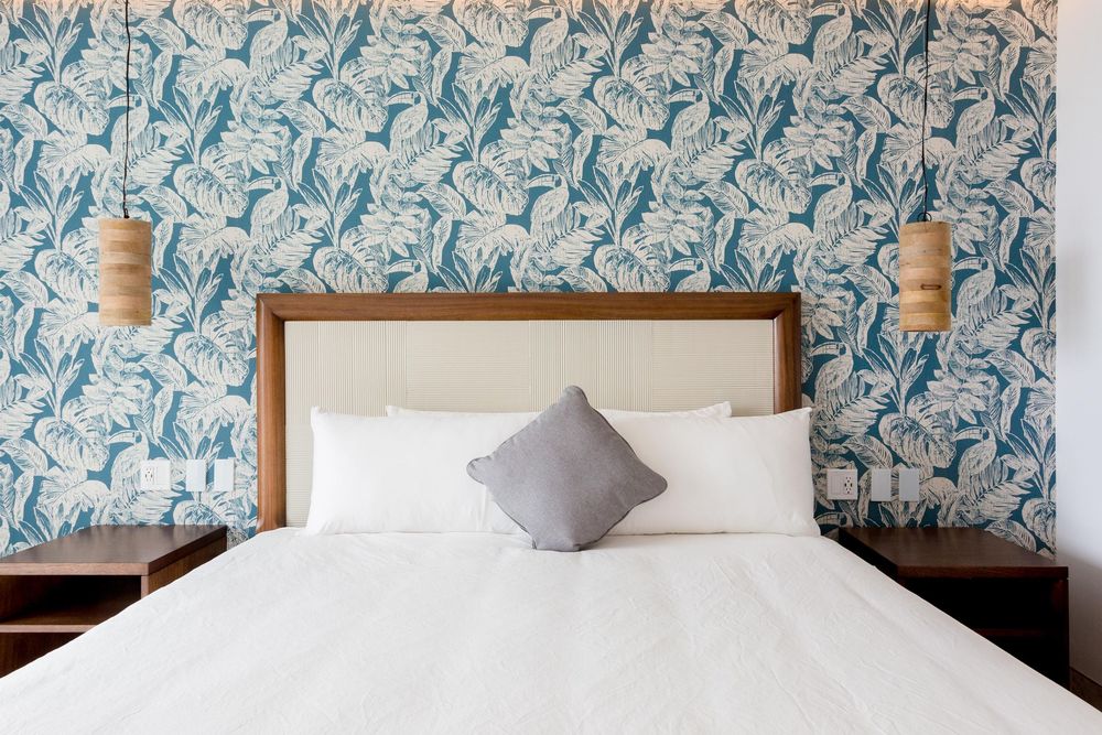 Hotel bed with white bedding and gray pillow against a blue tropical floral wallpaper with pendant lights.