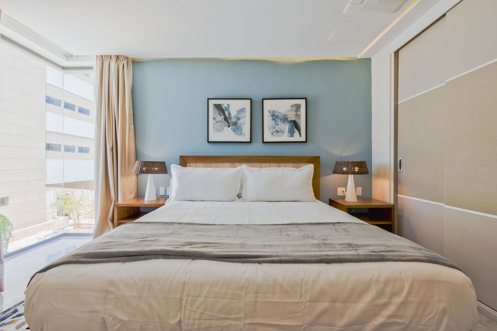 Modern hotel bedroom with blue accent wall, king bed, neutral linens, bird artwork, and natural light from large windows.