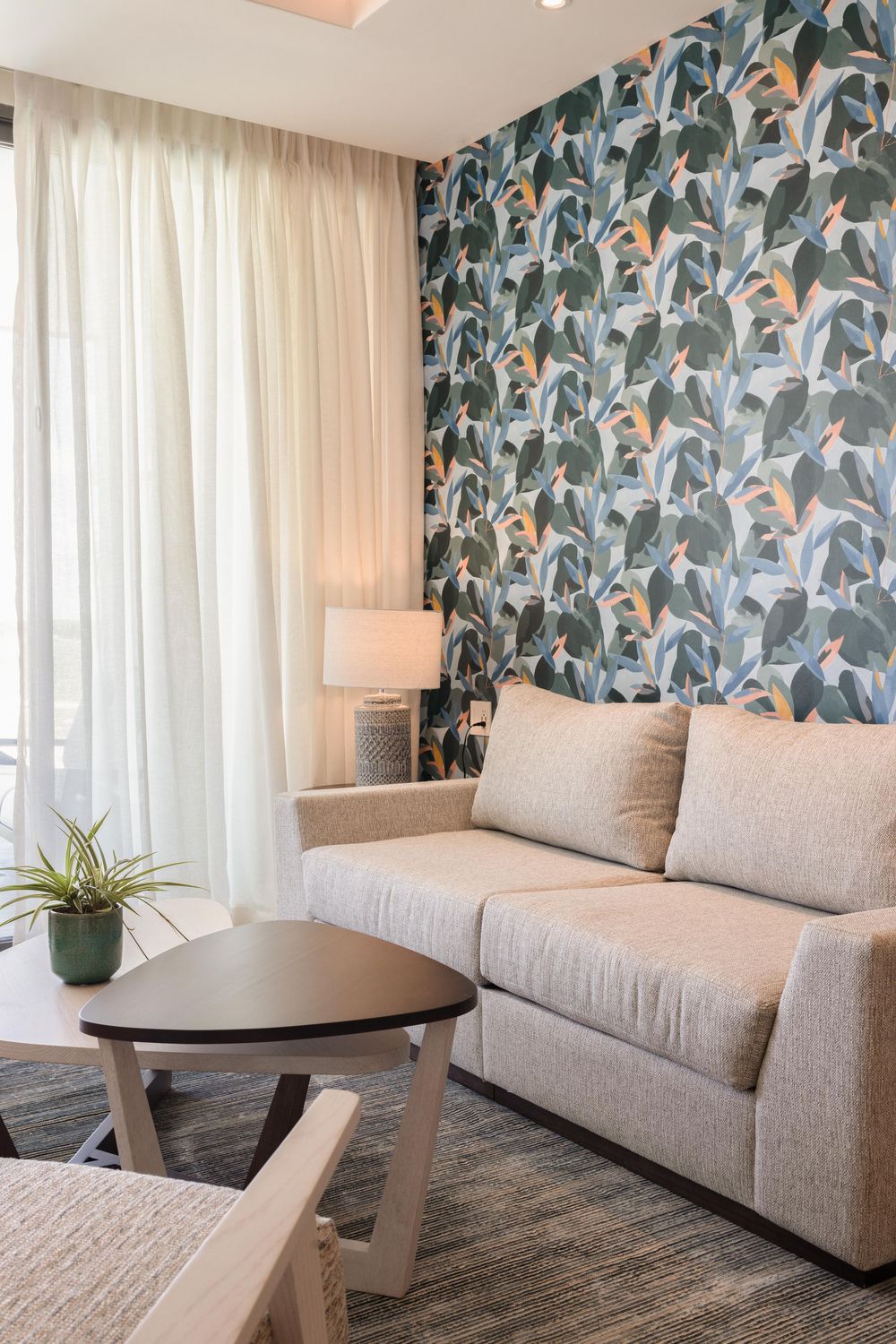 Modern hotel suite living area featuring a beige sofa, botanical wallpaper accent wall, and small round coffee table.