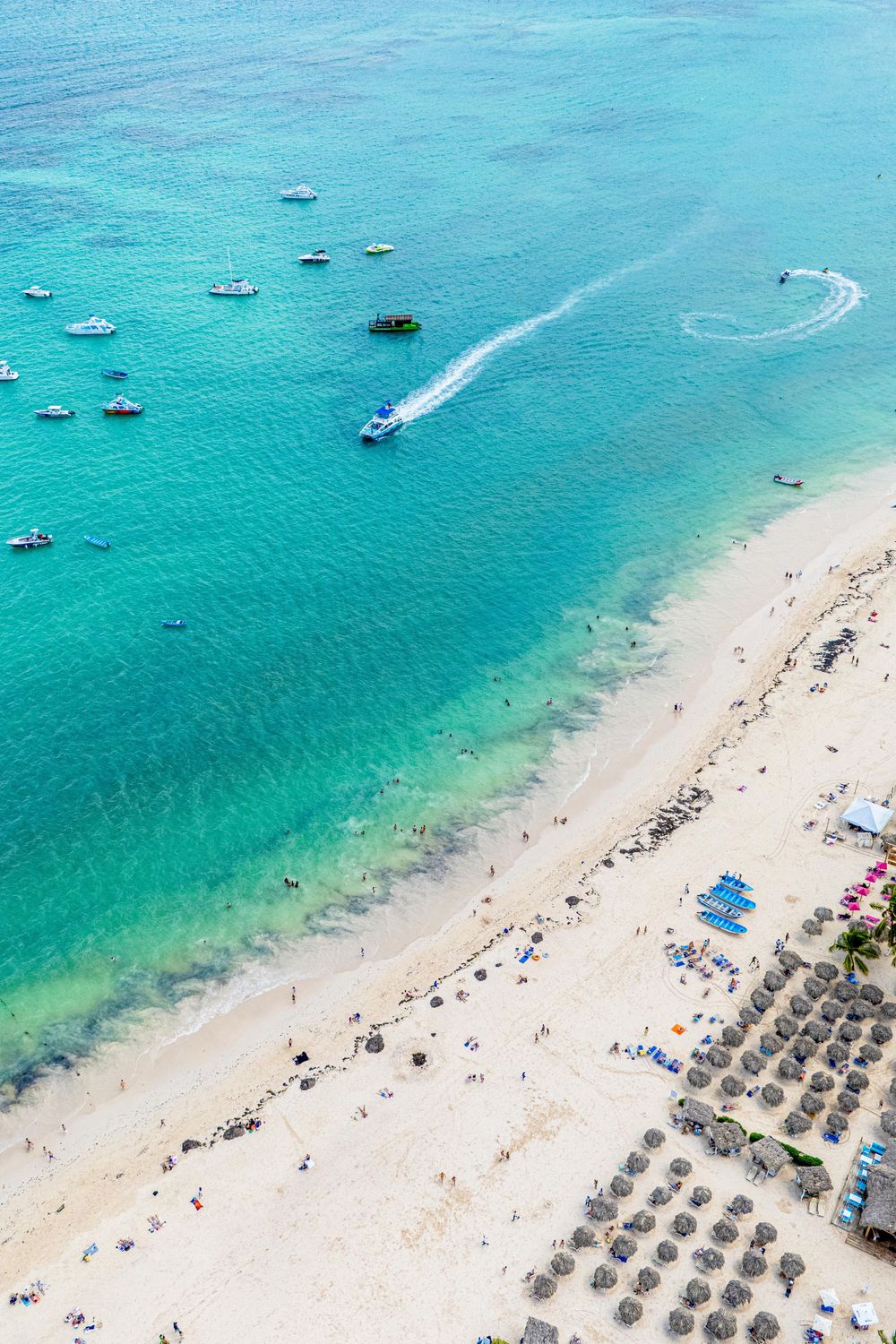 Aerial view of a pristine beach with turquoise waters, boats anchored offshore, and rows of beach umbrellas along the shore. Bavaro Beach