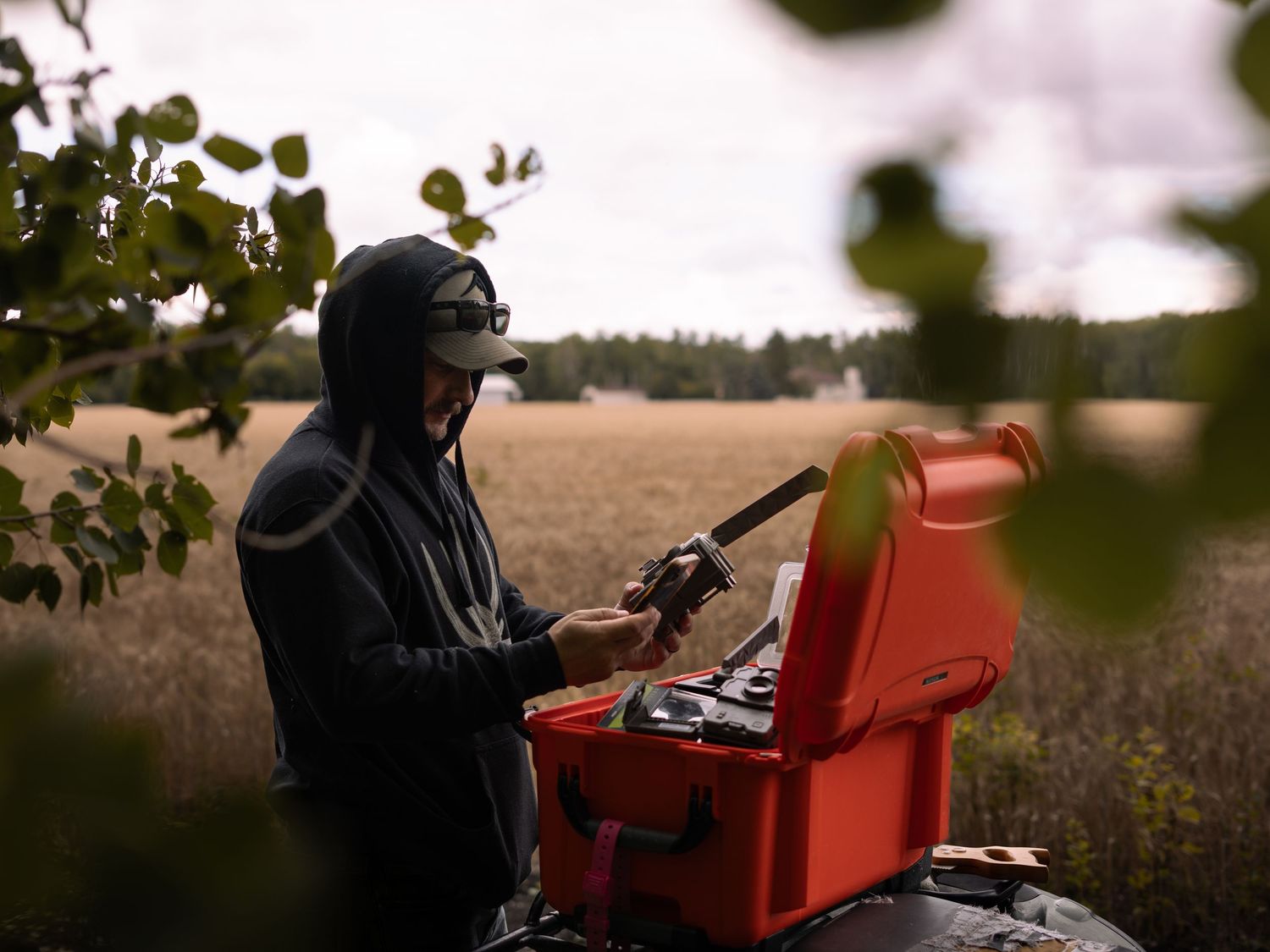 Person in dark clothing operating equipment with a red case in an outdoor field setting at dusk.