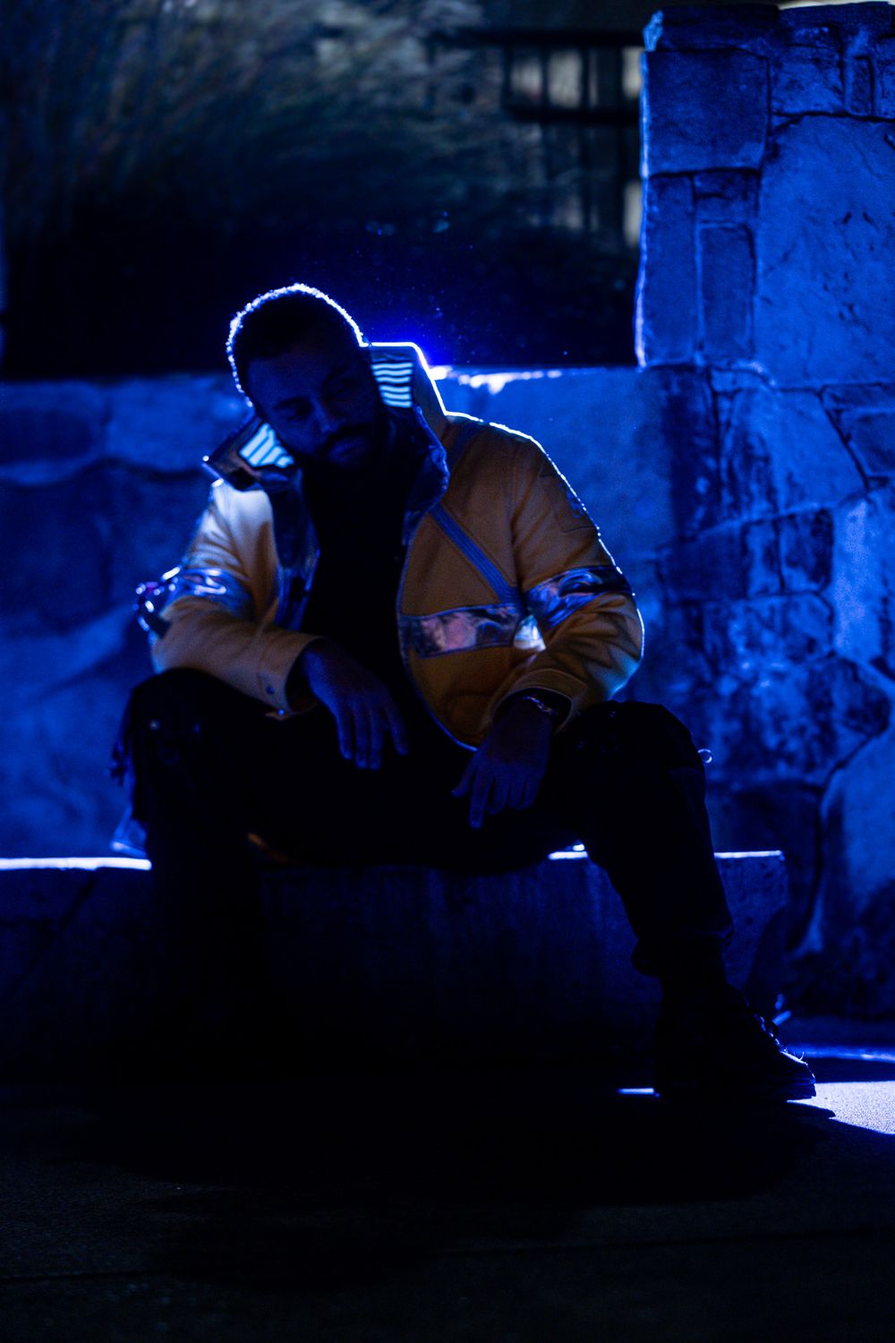 A silhouetted figure sits alone in moody blue lighting against a dark urban backdrop.