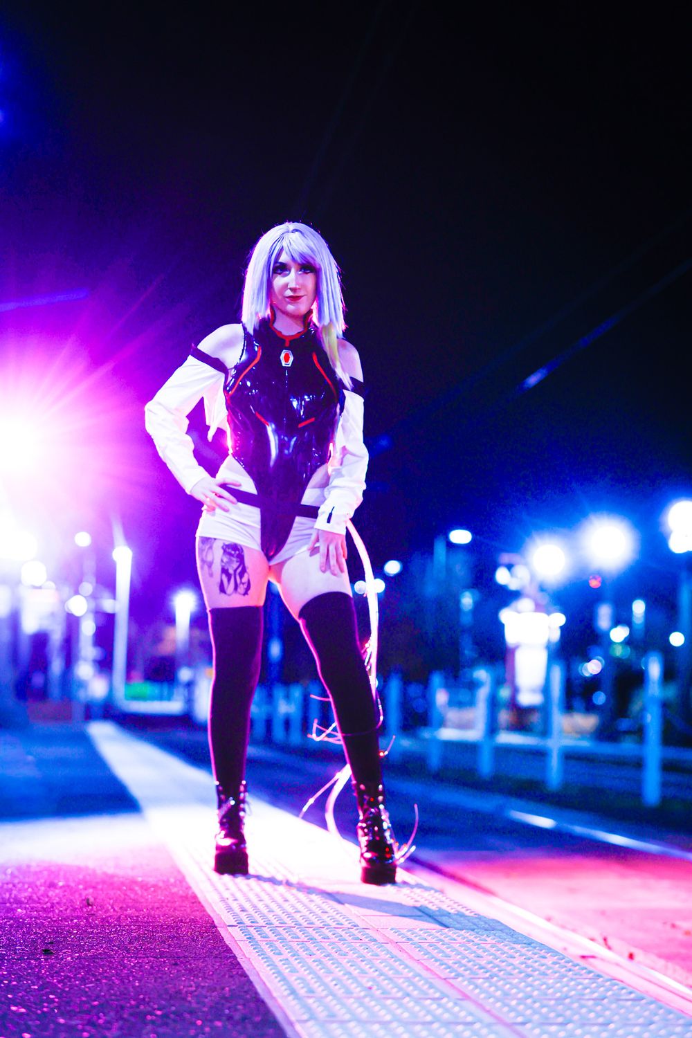 A figure in white and black clothing poses dramatically on an illuminated path with vibrant purple and blue lighting.