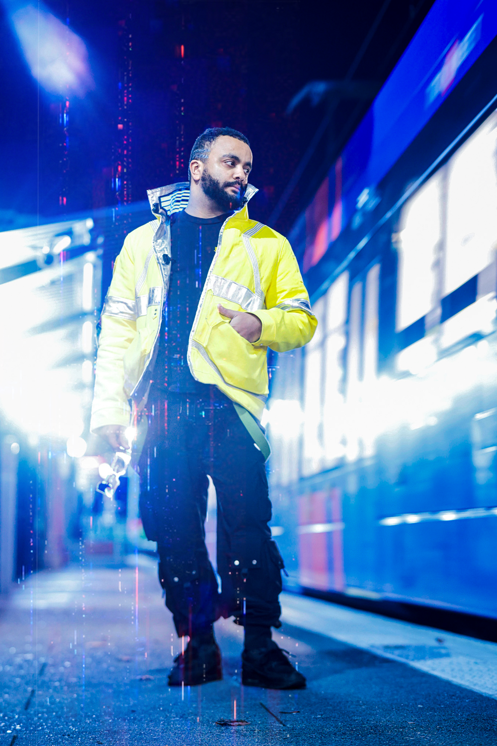 Person in yellow safety vest stands in dimly lit subway station with dramatic blue lighting and reflective surfaces.