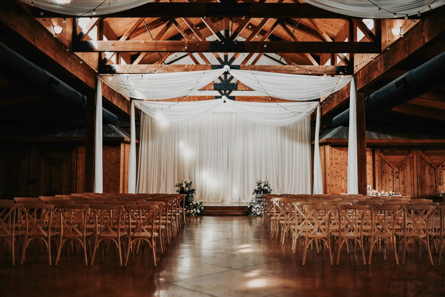 a romantic wedding ceremony site of wooden chairs lined in rows on a polished floor with twinkling edison lights hanging from the ceiling
