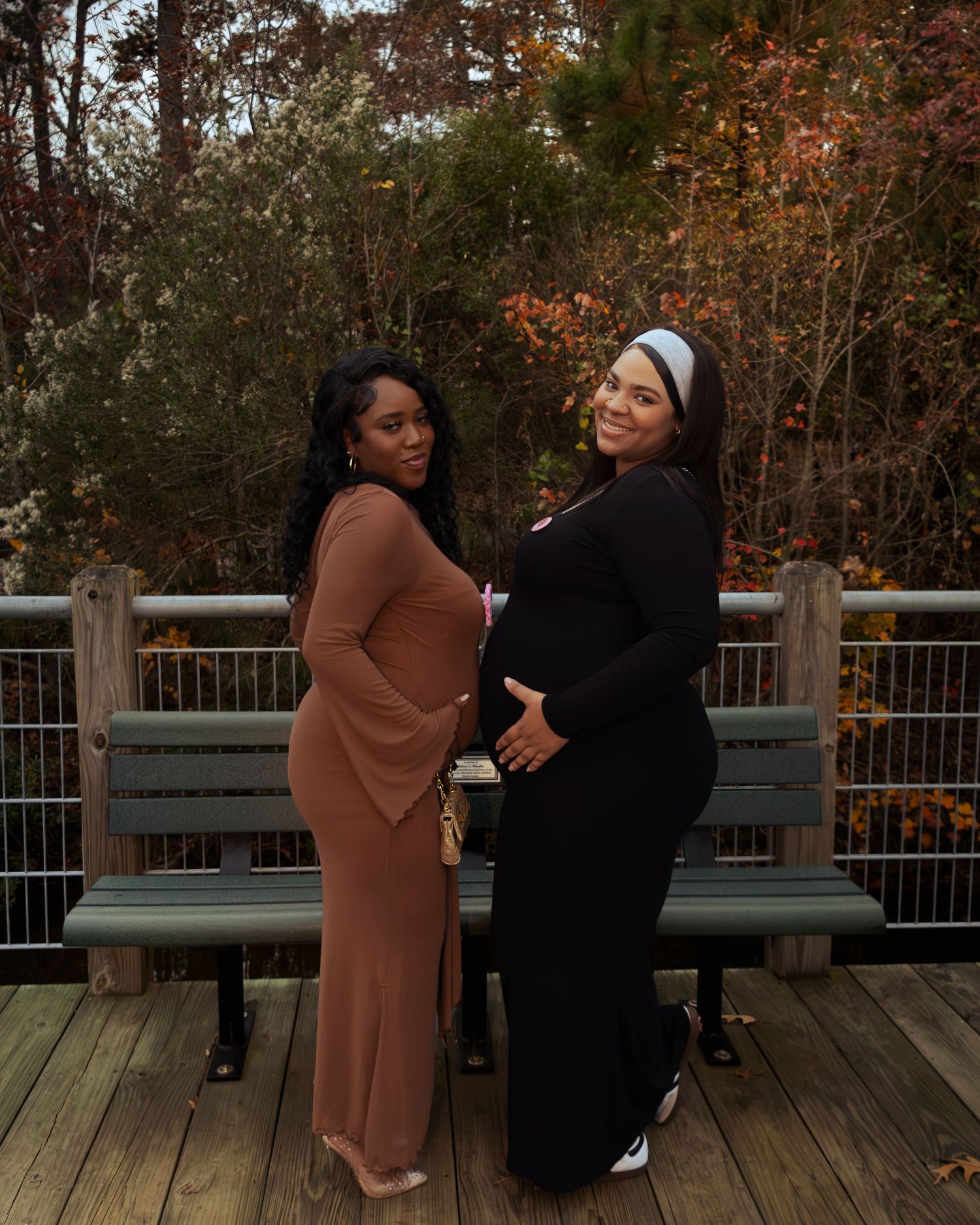 Two women in long dresses pose together on a wooden deck with autumn foliage in the background.
