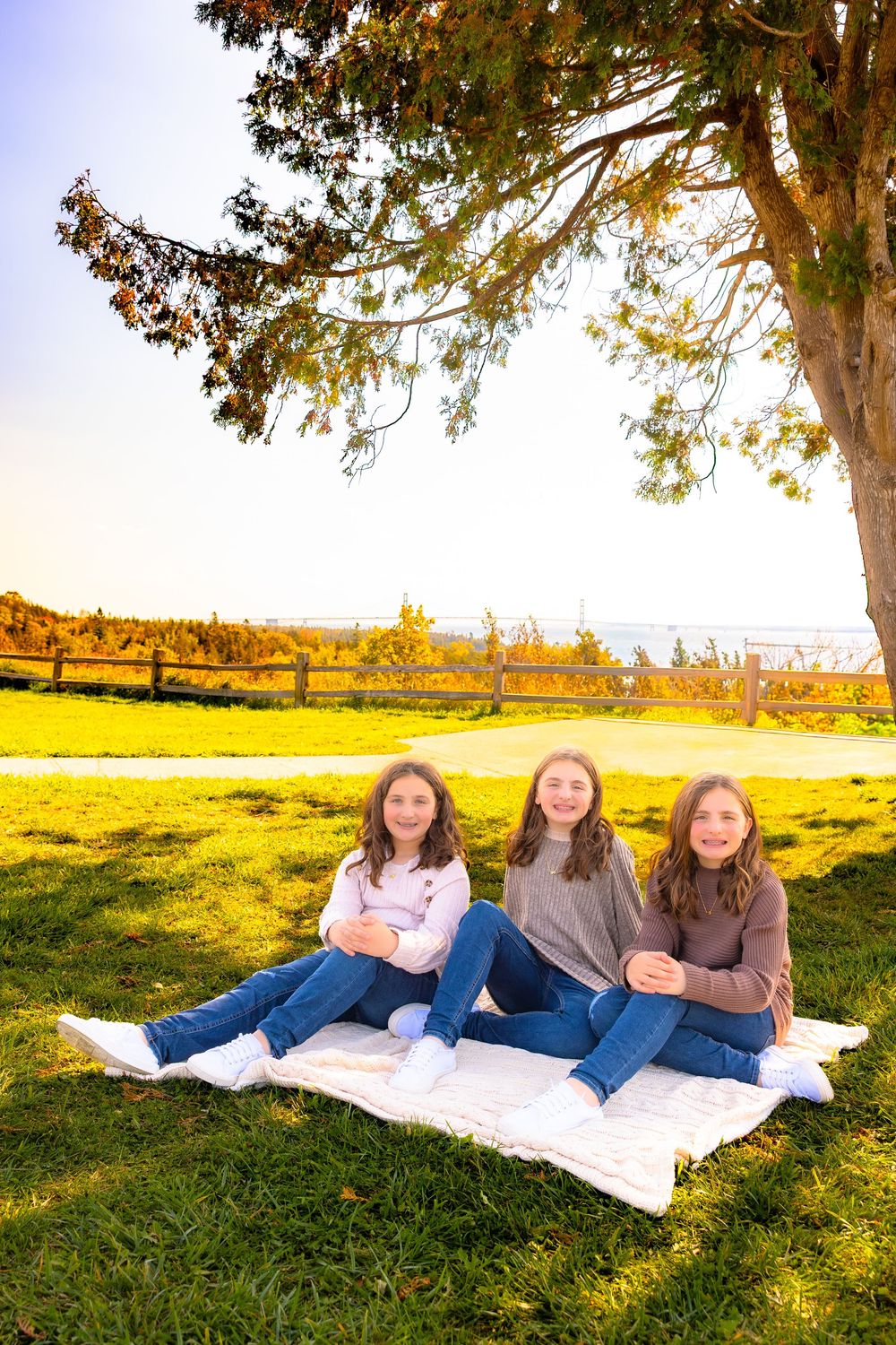 Three friends relax on a white blanket under a tree during golden hour in an autumn field with mountain views.