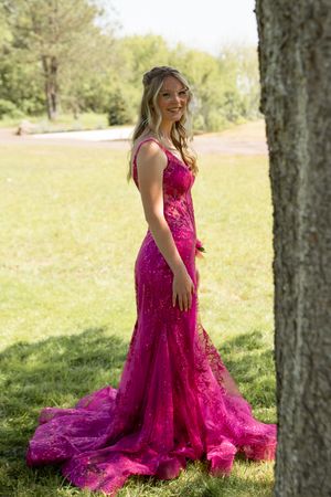 Person in a stunning magenta lace mermaid gown with long train poses outdoors in a sunlit garden setting.