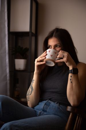 A cozy indoor scene shows someone in grey enjoying coffee near a window with houseplants in the background.