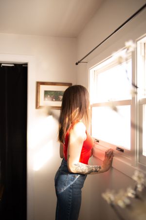 A person wearing a red tank top and jeans looks out a sunny window in a minimalist interior setting.