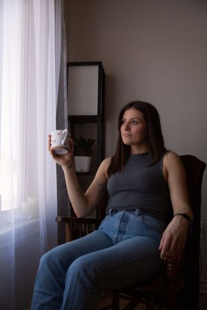 Someone wearing a grey tank top and jeans relaxes in a chair by a window while holding a coffee cup.