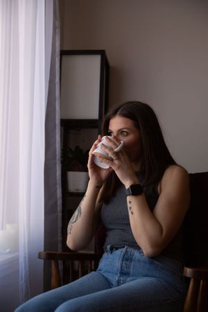 Person in casual attire sips from a white mug while seated in a wooden chair near a window.