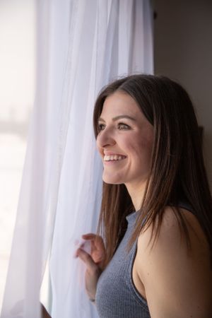 Person in sleeveless top smiles brightly while standing near a sunlit window with sheer curtains.