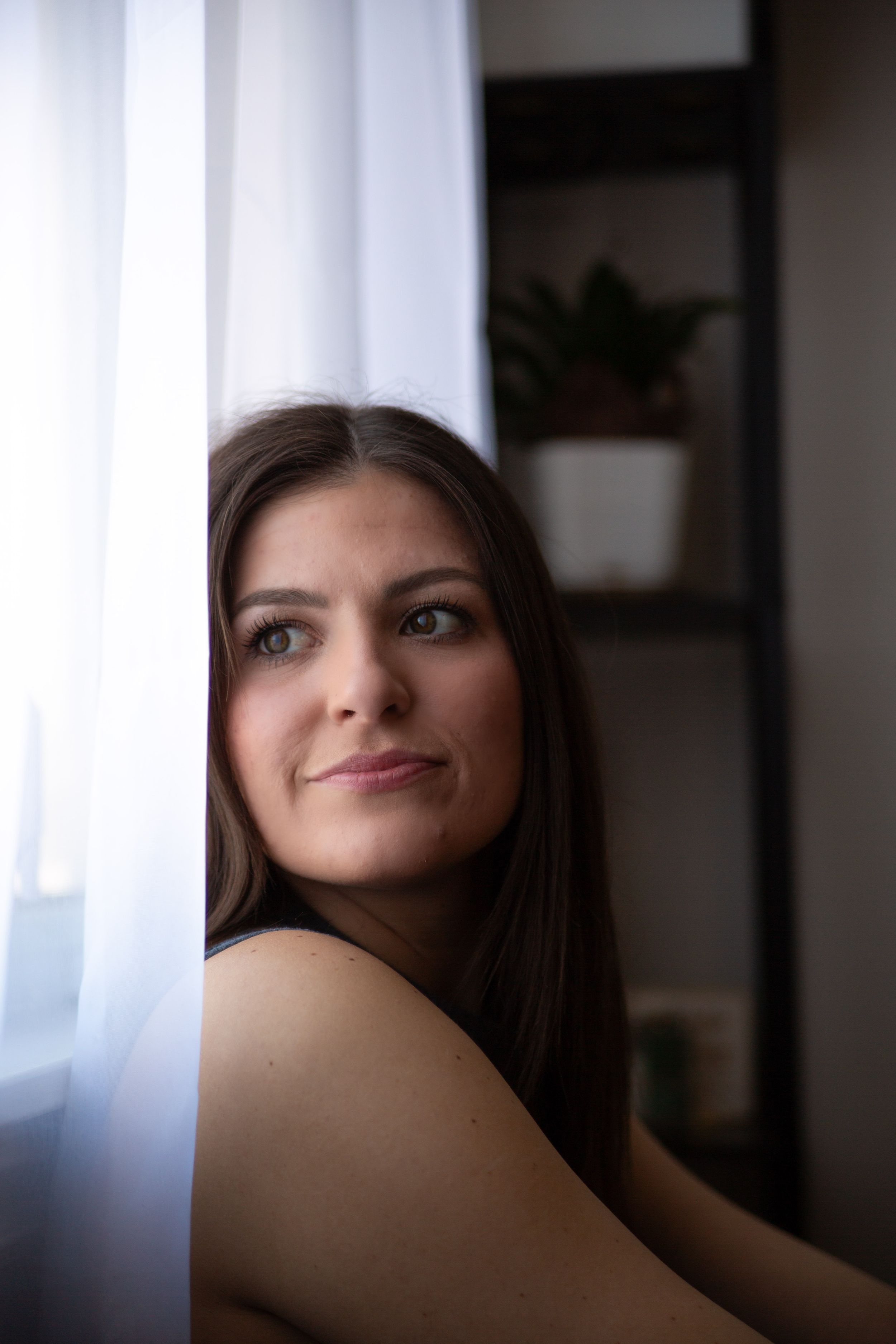 A soft natural light portrait shows someone gazing thoughtfully by a window with sheer curtains.