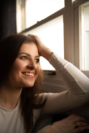 Figure in light clothing smiles while relaxing by a bright window in natural lighting.