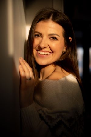 A smiling person with long brown hair poses in warm lighting wearing a gray off-shoulder top.
