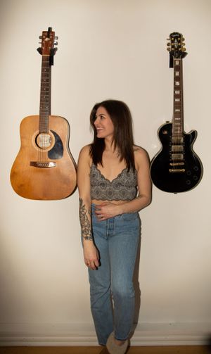 An acoustic and electric guitar flank someone in a gray top and jeans standing against a white wall.
