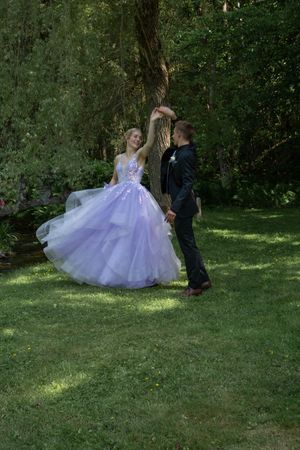 Two figures in formal attire dance gracefully on a lush green lawn surrounded by trees in a romantic outdoor setting.