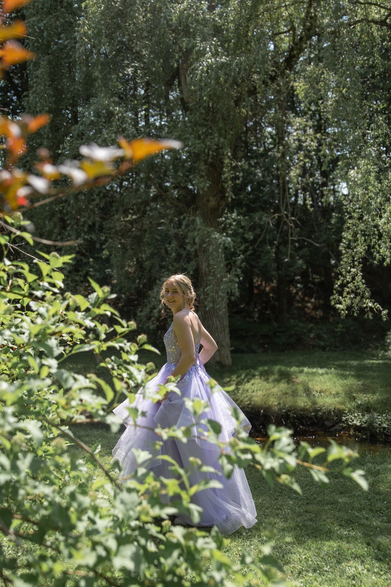 A figure in a flowing white dress twirls gracefully in a sunlit garden surrounded by lush greenery and trees.