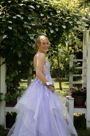 Person in a flowing lavender ballgown poses gracefully on a porch surrounded by lush green foliage.