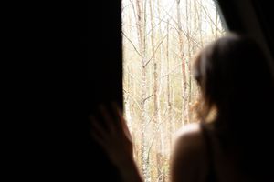 A silhouetted figure stands in front of a bright window with sunlight streaming through bare trees outside.