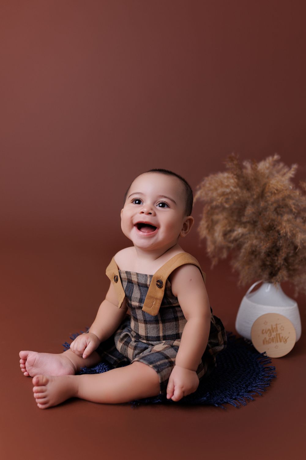Smiling baby in plaid overalls sits on brown backdrop with decorative pampas grass.