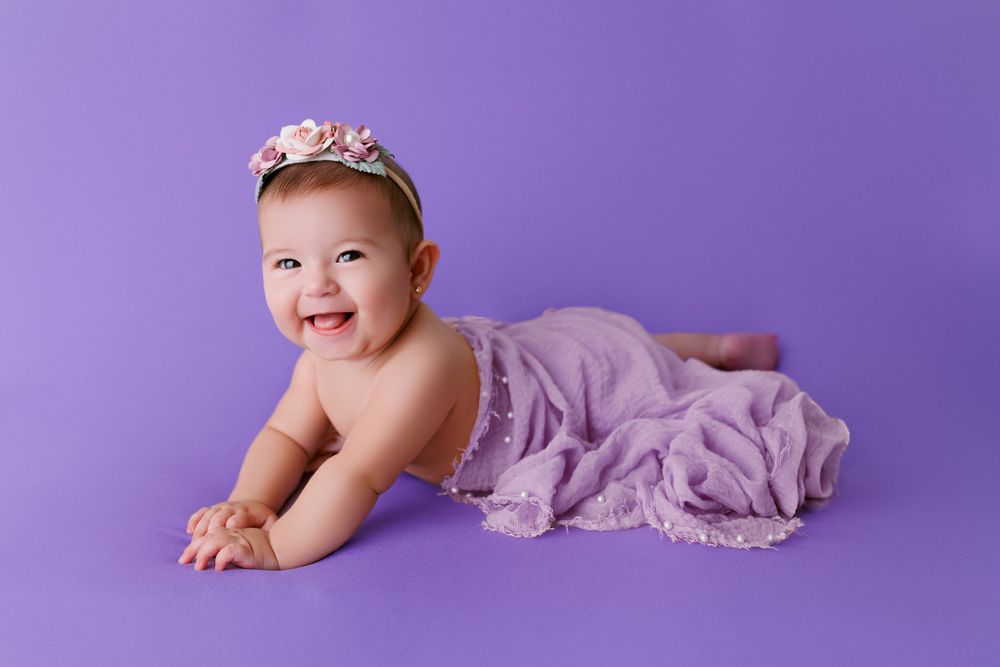 Baby lies on purple backdrop during milestone photo session.