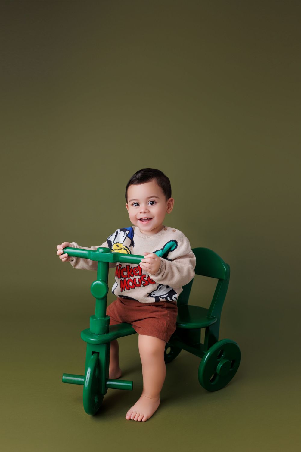 Baby sits in vintage green tractor prop against olive backdrop during professional photo session.