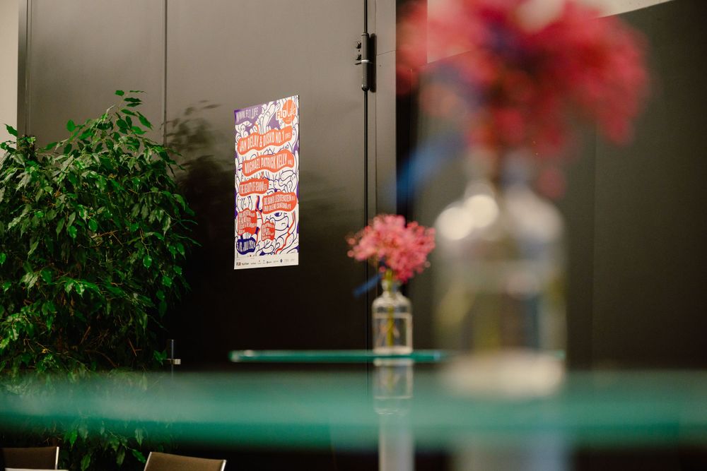 Pink flower in vase with poster on dark wall in background.