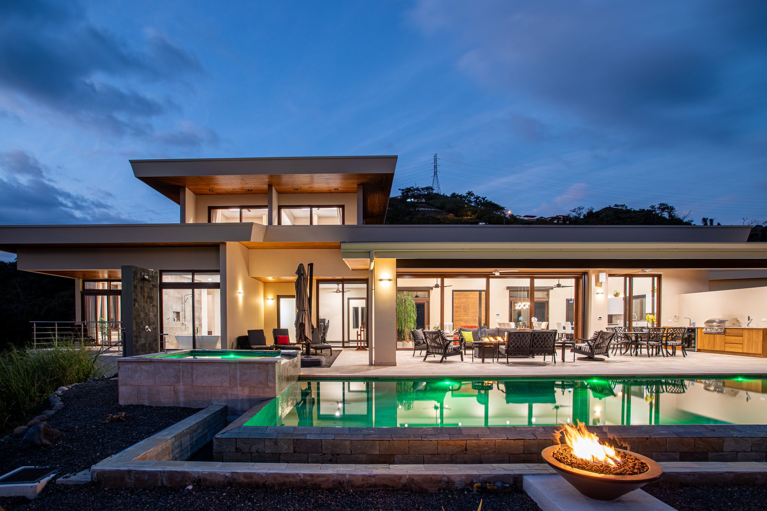 Modern luxury home at dusk with illuminated swimming pool and fire pit, featuring contemporary architecture and outdoor seating area