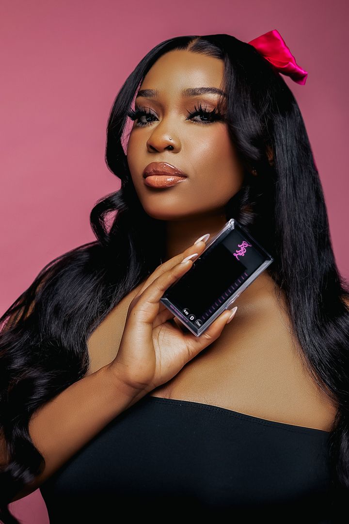 A beauty portrait with long dark hair and a red bow against a pink background holding a product.