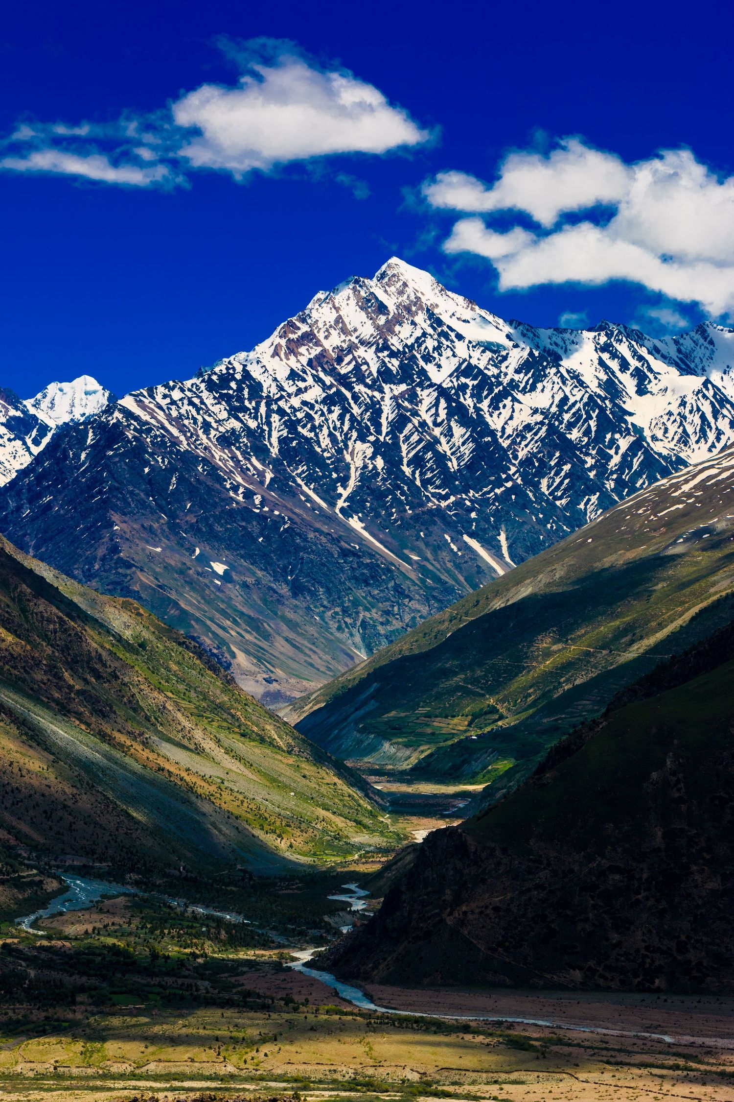 Snow-capped mountain peak rises above a winding valley with rivers and green slopes under blue skies.