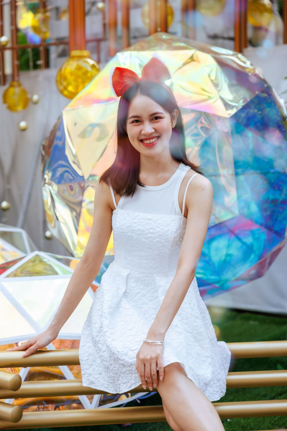 Person in white dress sits on bamboo seating against iridescent backdrop wearing red bow accessory.