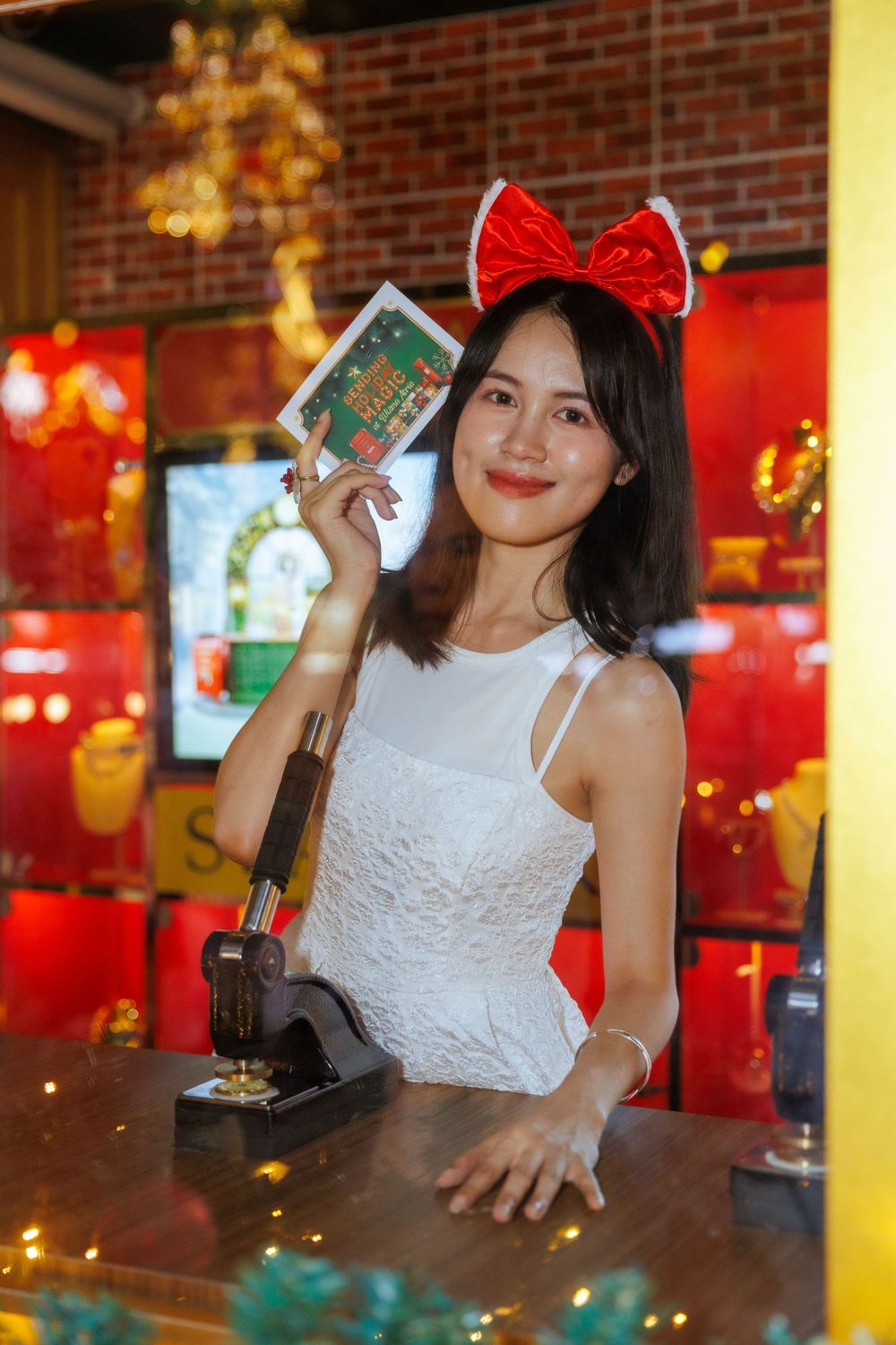 Person in white lace dress at counter holding up card with red bow headband in decorated shop.