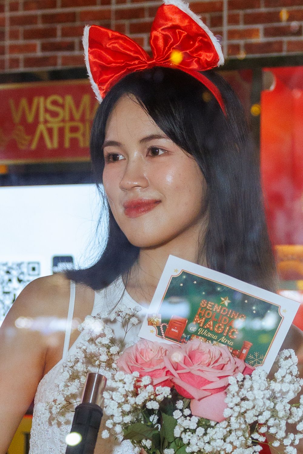 Person in white dress holding book and flowers with red bow against brick wall and holiday decorations.