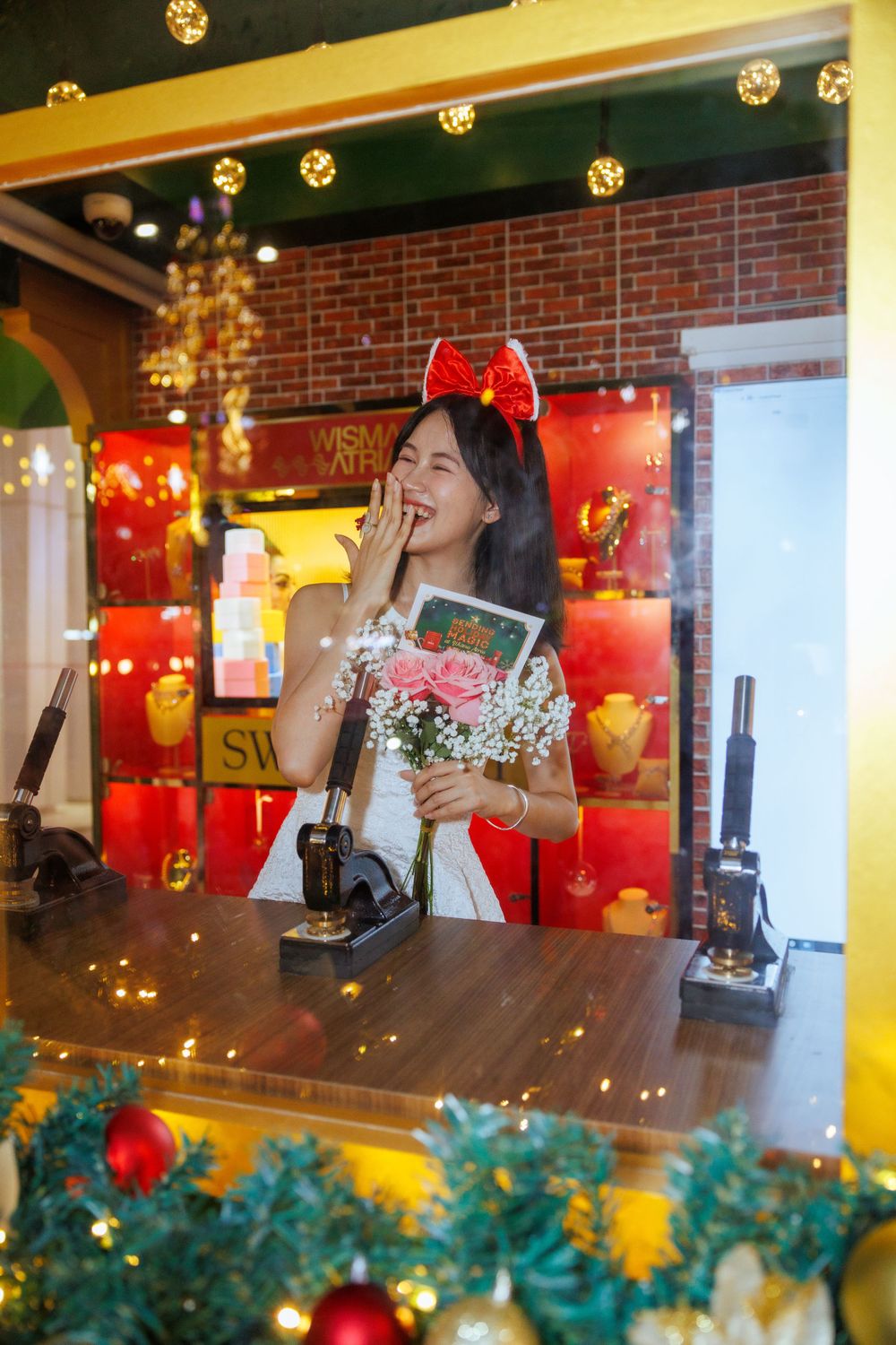 Person at counter wearing white dress and red bow headband, holding flowers and card with festive lights.