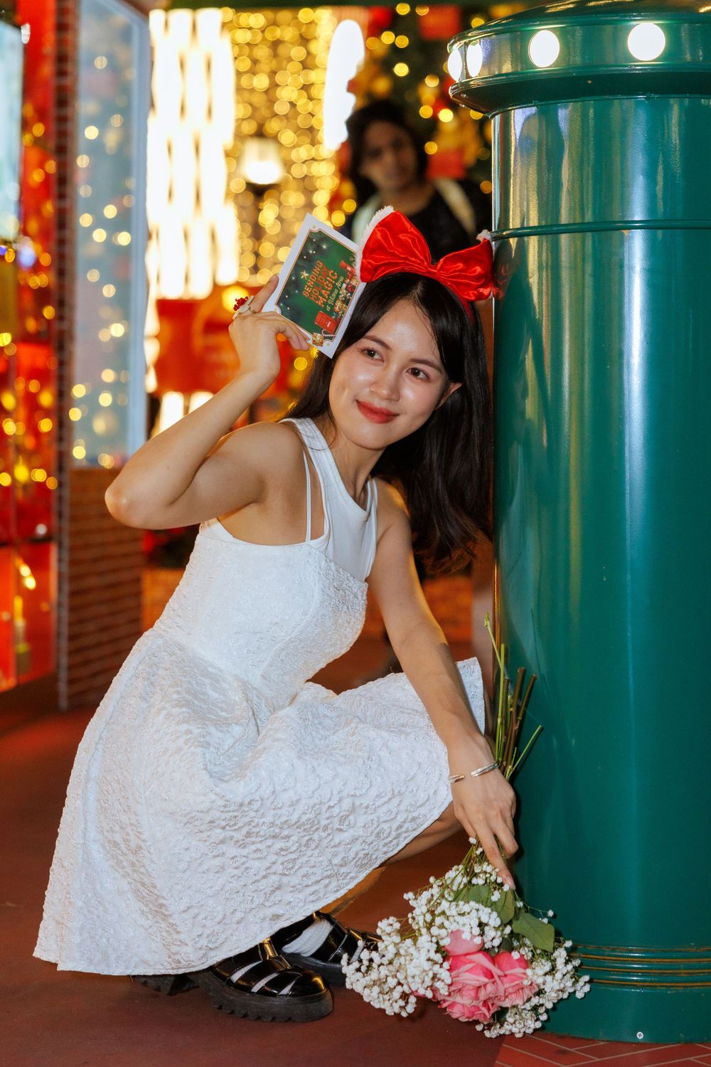 Person in white dress crouching by green pillar with flowers and card, wearing red bow.