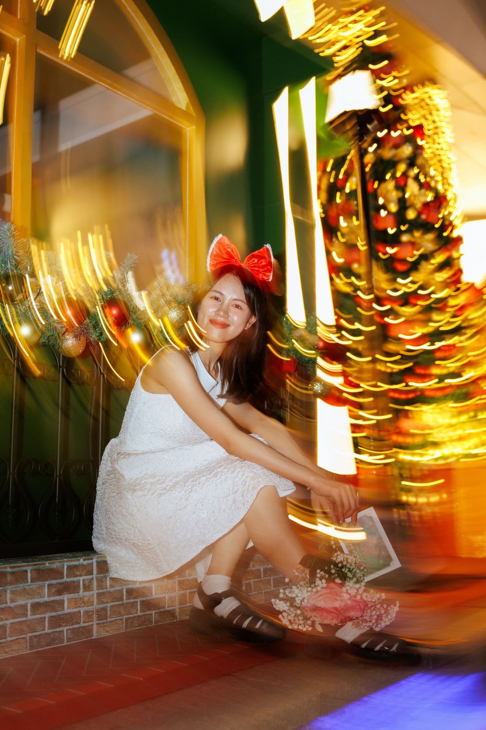 Person in white dress posing with flowers against blurred holiday lights and decorations.