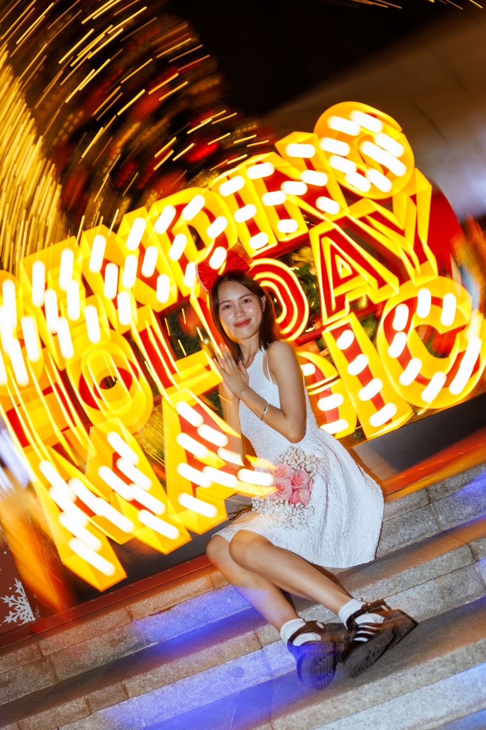 Person in white dress sitting on steps with swirling neon lights and holiday decorations in background.